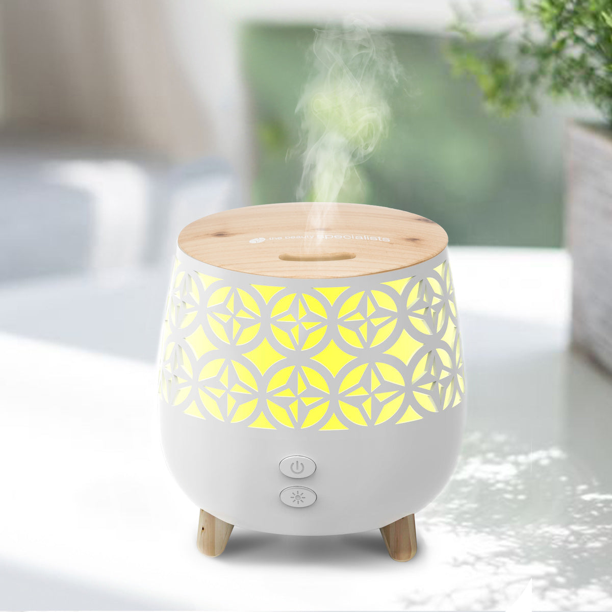 The Rio TALIA Aroma Diffuser, Humidifier & Night-Light features a wooden top, yellow glow, and geometric cutouts, releasing soothing vapor on a white surface with a blurred background.