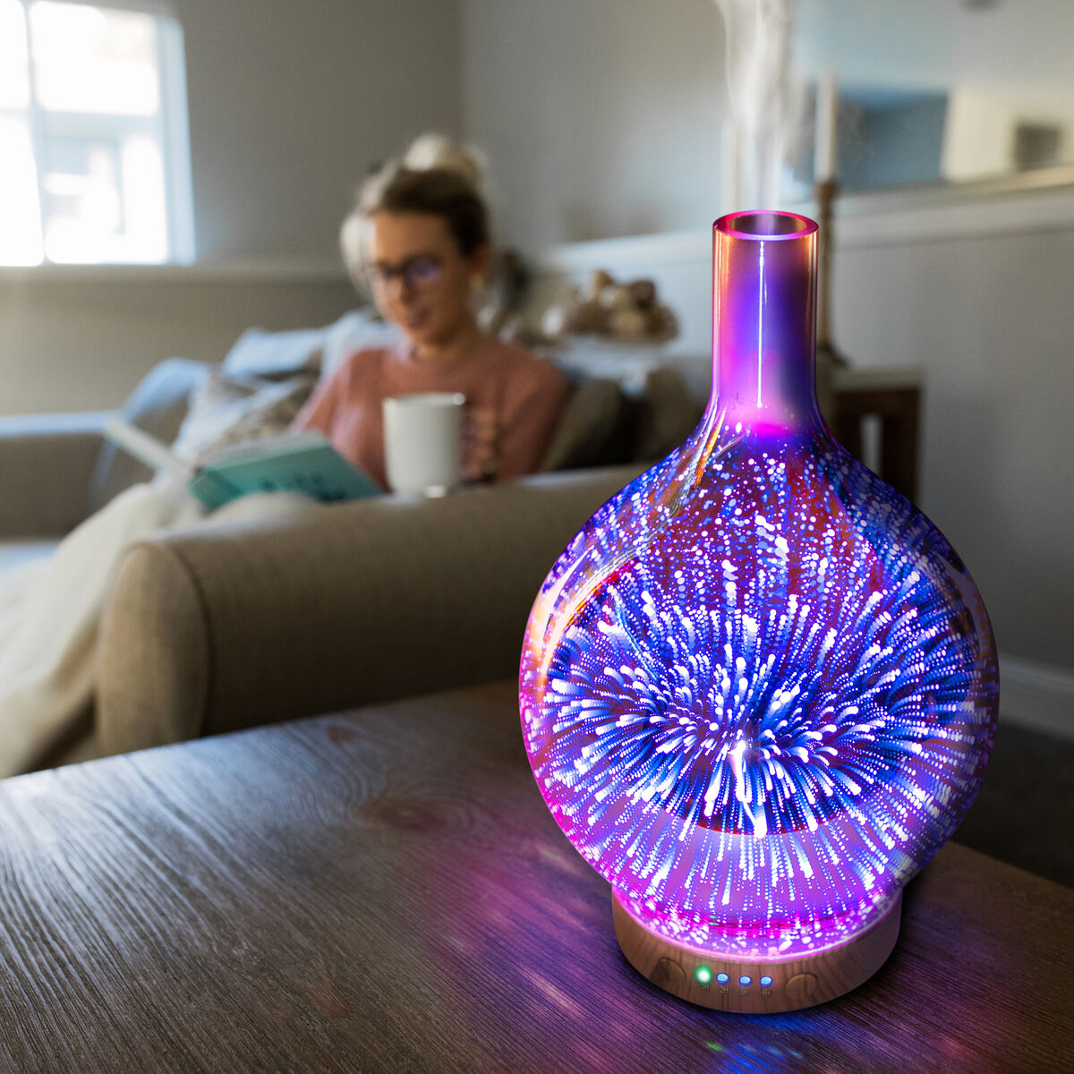The Rio ELLA Aroma Diffuser, Humidifier and Night-Light emits a soothing mist on a wooden table as someone reads with a mug in hand in the softly lit living room, creating a cozy and relaxing atmosphere.