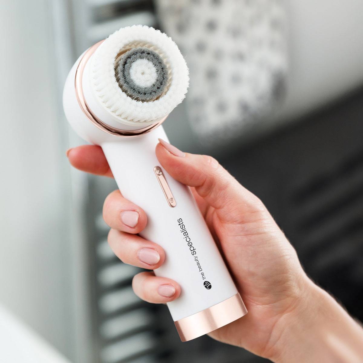 A hand holds the Rio the Beauty Specialists PURE CLEANSE Deep Cleansing & Exfoliating Facial Brush with a rotating bristle head, ideal for deep skincare routines, against a blurred bathroom background.