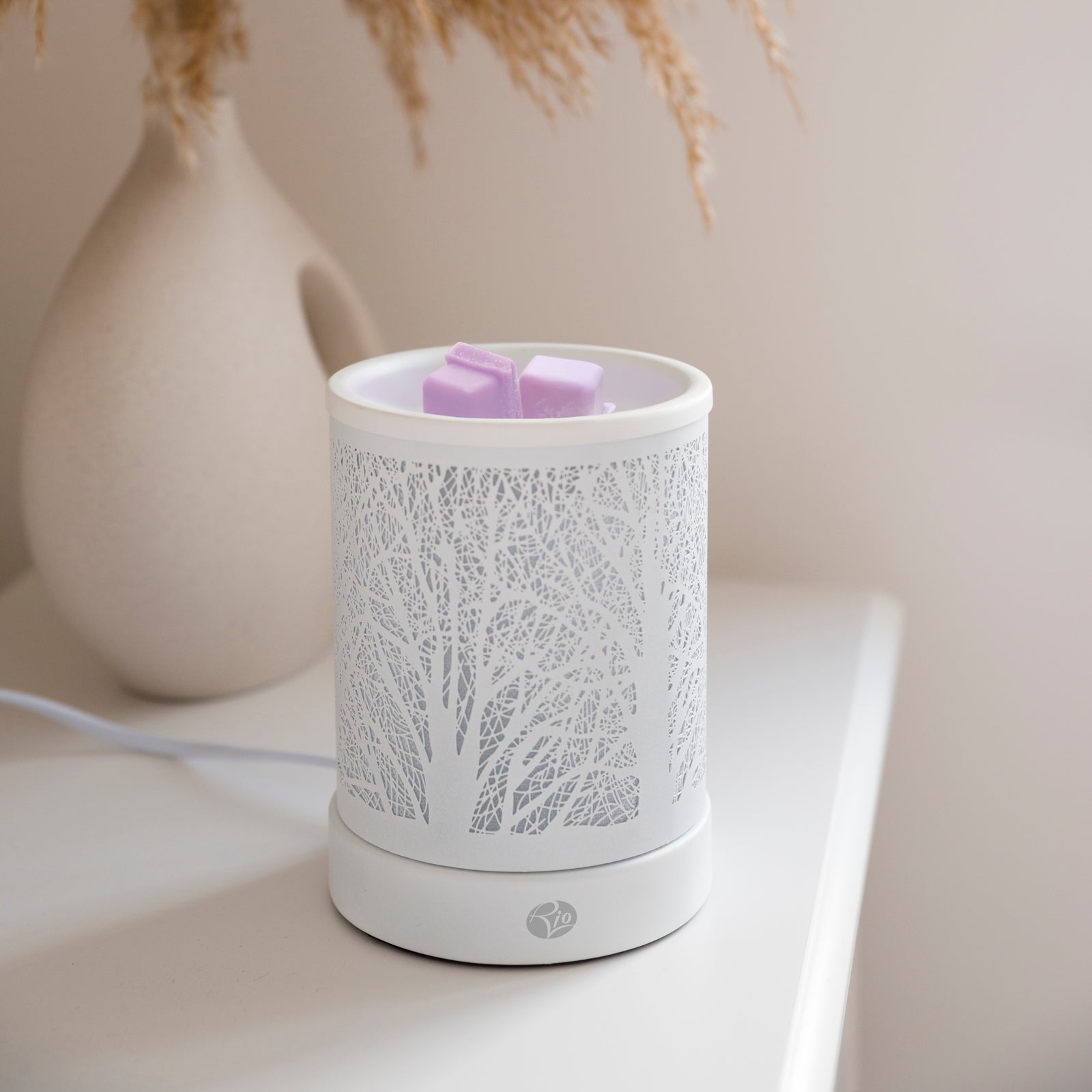 The Rio FORA Wax Melt Burner & Aroma Diffuser Lamp, featuring a tree branch pattern, melts two purple wax cubes on a white surface. A beige vase with dried grass in the blurred background adds to the relaxing ambiance.