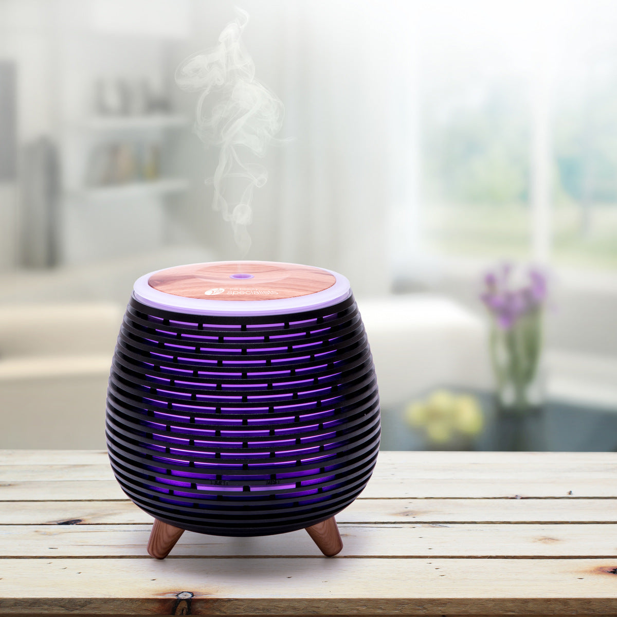 The Rio ZOEY Aroma Diffuser, Humidifier & Night-Light in black with wood grain emits a purple glow and soothing mist on a wooden table in a bright living room, creating a peaceful atmosphere for relaxation.