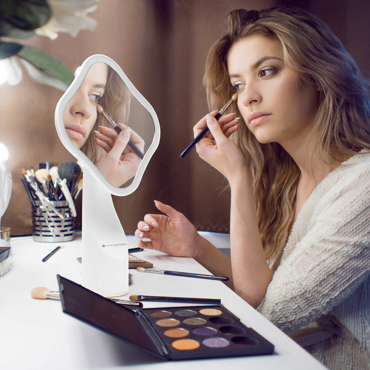 A woman sits at a vanity applying eyeshadow with a brush while looking into the Rio Illuminated Full Size Beauty Mirror. Makeup brushes and a vibrant eyeshadow palette are arranged on the table in front of her.