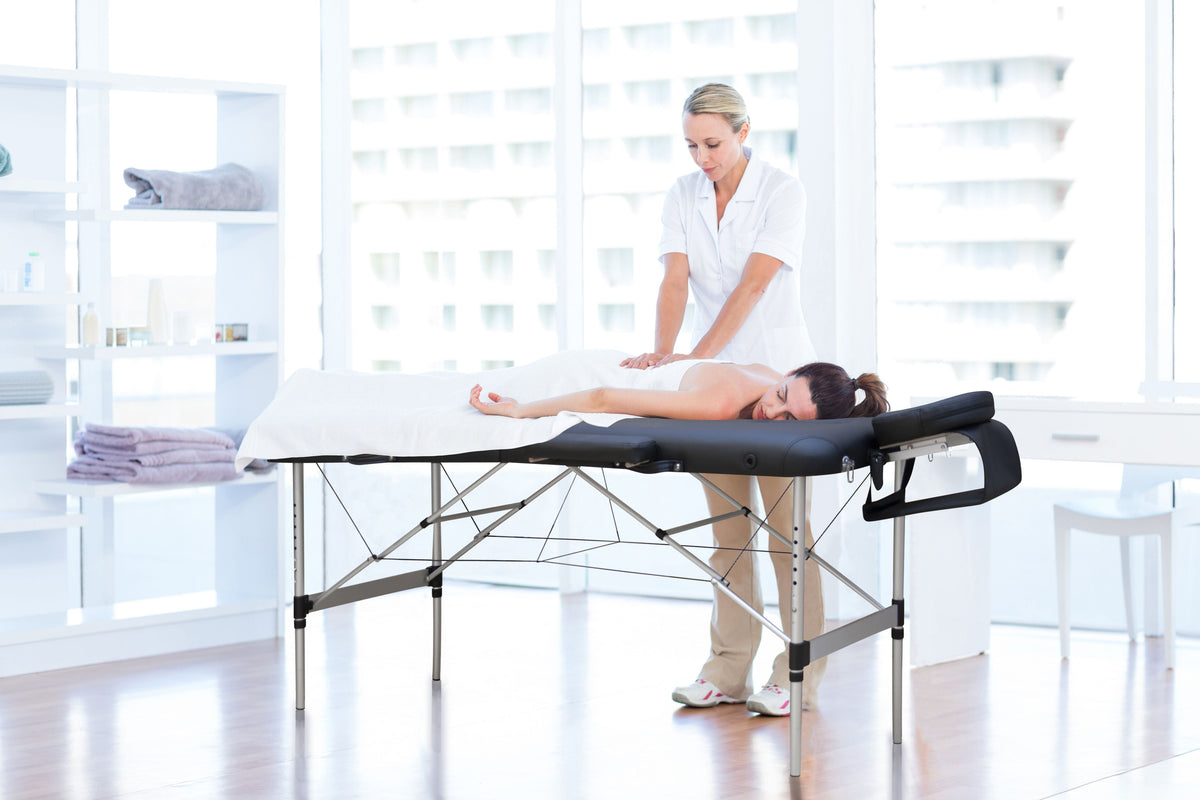 A woman relaxes face down on the Rio Professional Aluminium Massage Table & Treatment Couch while a therapist in white works on her back in a bright, modern room with large windows.