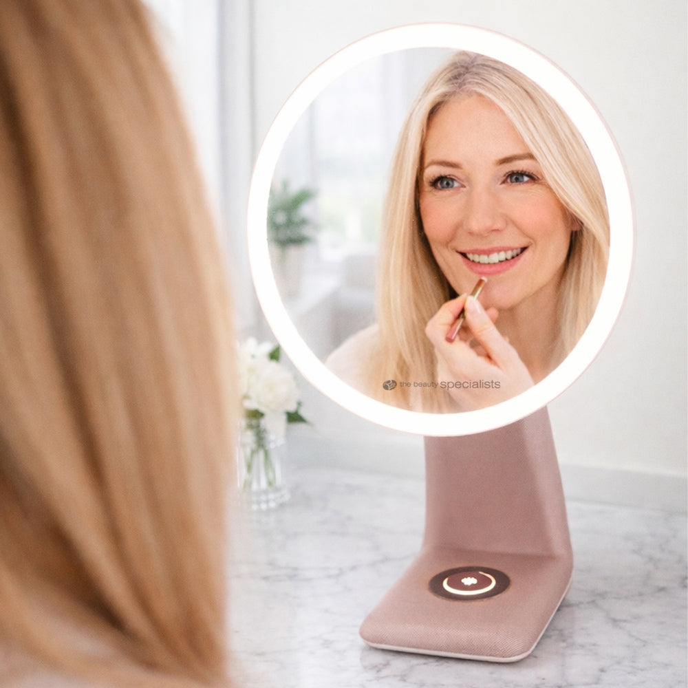 A woman with long blonde hair smiles as she applies lipstick, looking into the Rio TrueColour High Performance Makeup Mirror—a round, illuminated vanity mirror with high clarity and a convenient touch control on its base.