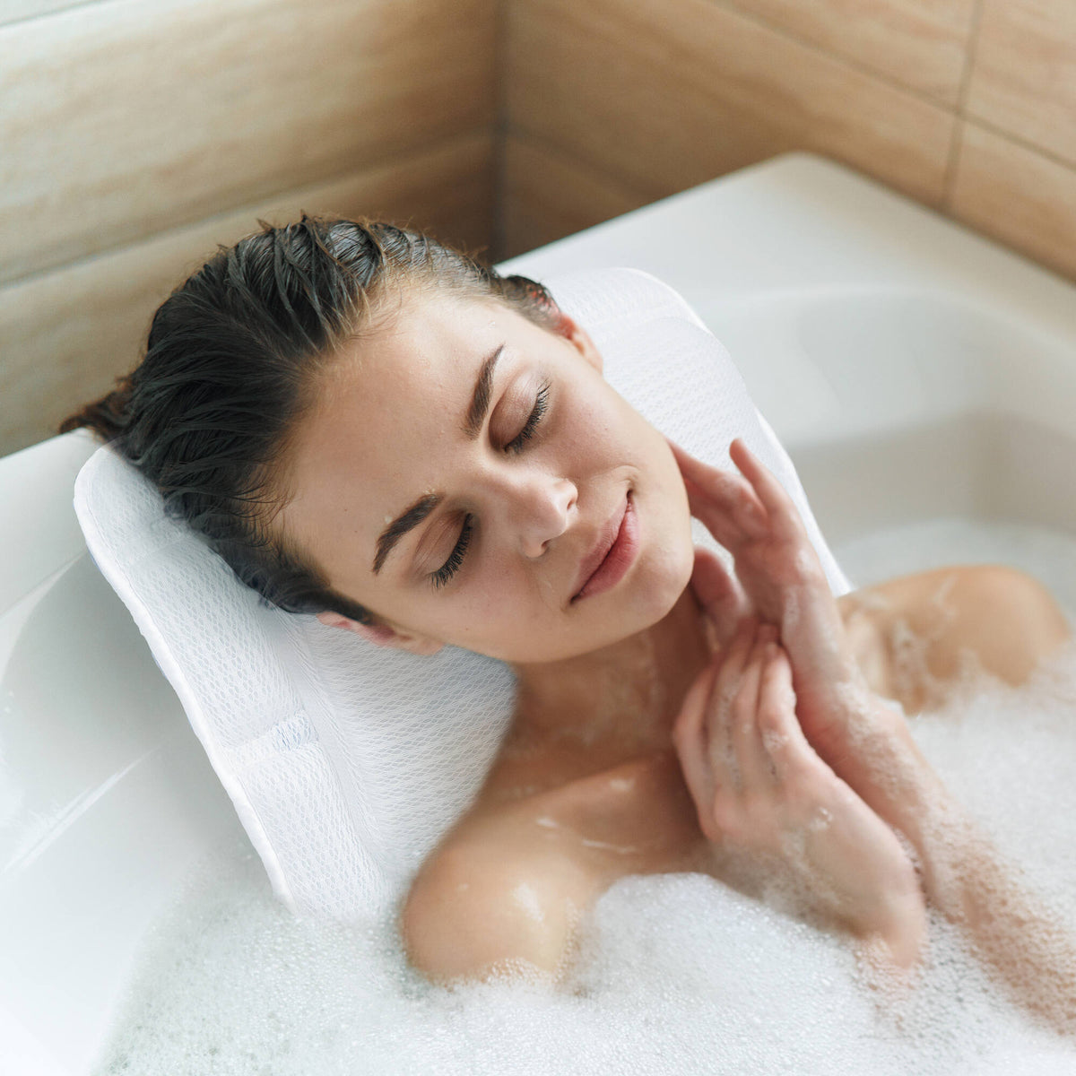 A woman with closed eyes relaxes in a bubble bath, resting her head on the Rio Luxury Bath Mat & Pillow, gently touching her neck and enjoying a moment of true bathing indulgence amid white tiles.