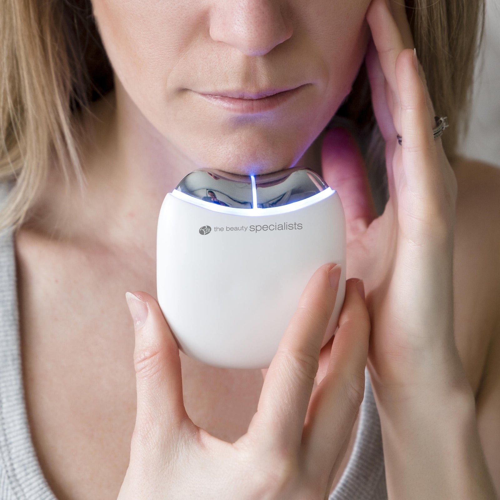 A woman in a gray tank top uses the Rio Face Lift & Tone Facial Toner, holding the white, curved device with a glowing blue light to her chin for skin toning and anti-aging benefits.