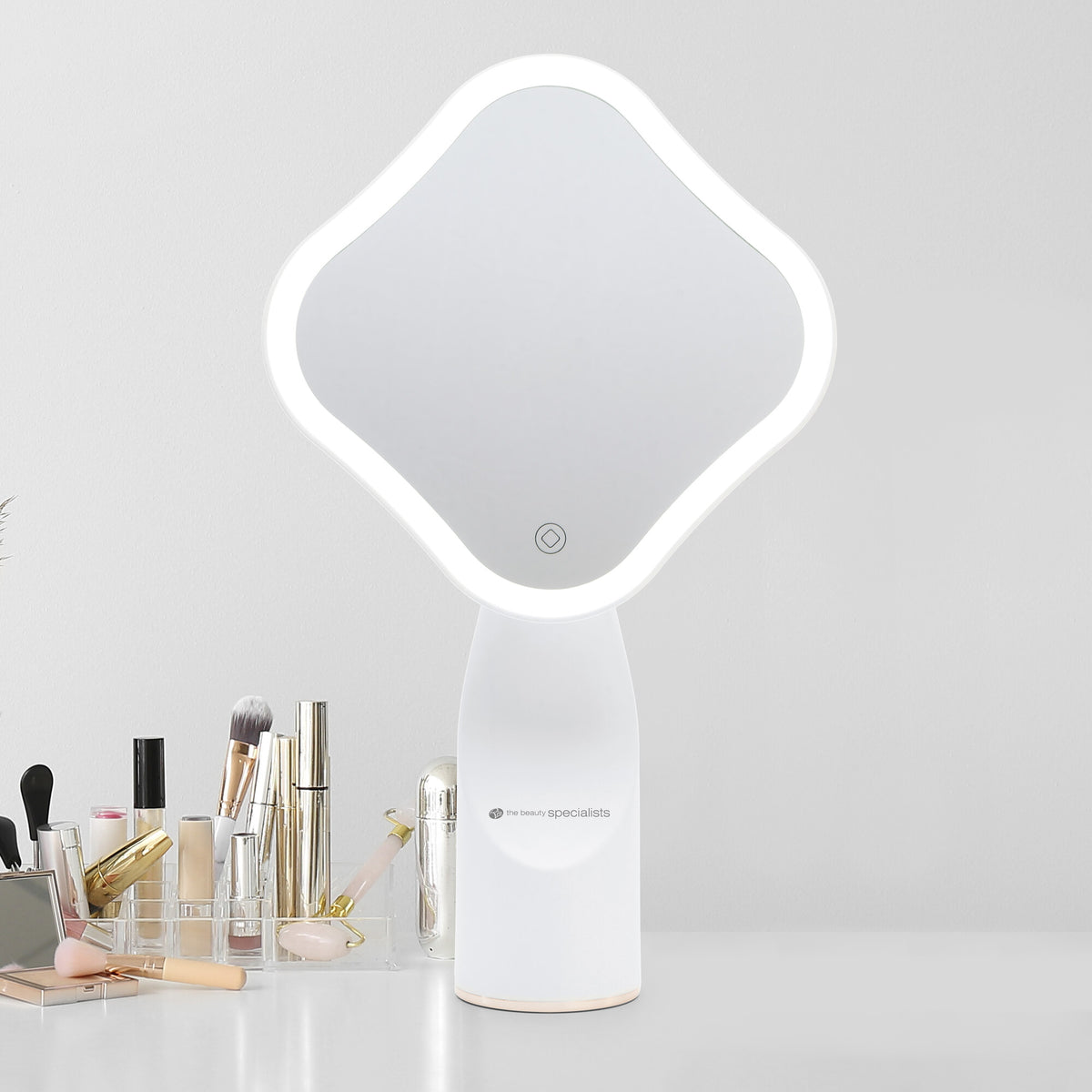 The Rio Illuminated Full Size Beauty Mirror with a white frame sits on a white table, surrounded by neatly arranged makeup brushes, cosmetics, and beauty products against a plain light gray wall.