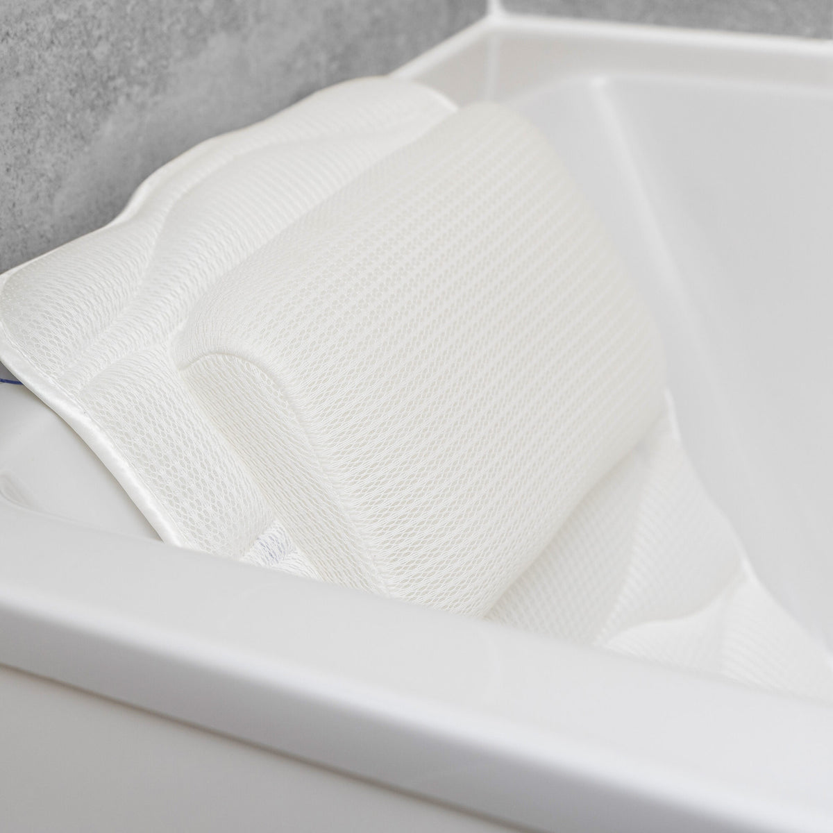The Rio Luxury Bath Mat & Pillow, in white with a textured design, rests on a bathtub edge for head and neck support. Its contoured shape adds bathing comfort and relaxation, beautifully contrasted by a gray tiled background.