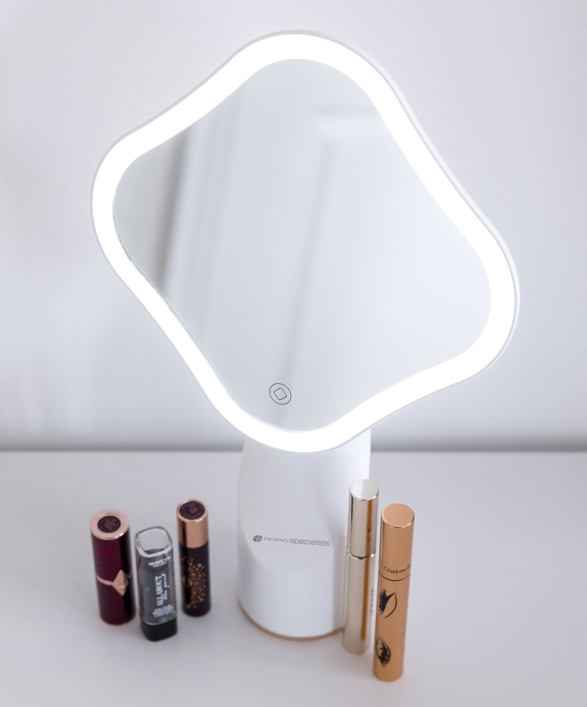 The Rio Illuminated Full Size Beauty Mirror, featuring a cordless wavy square design, sits on a white surface with five decorative makeup products in front. The background is plain and minimal.