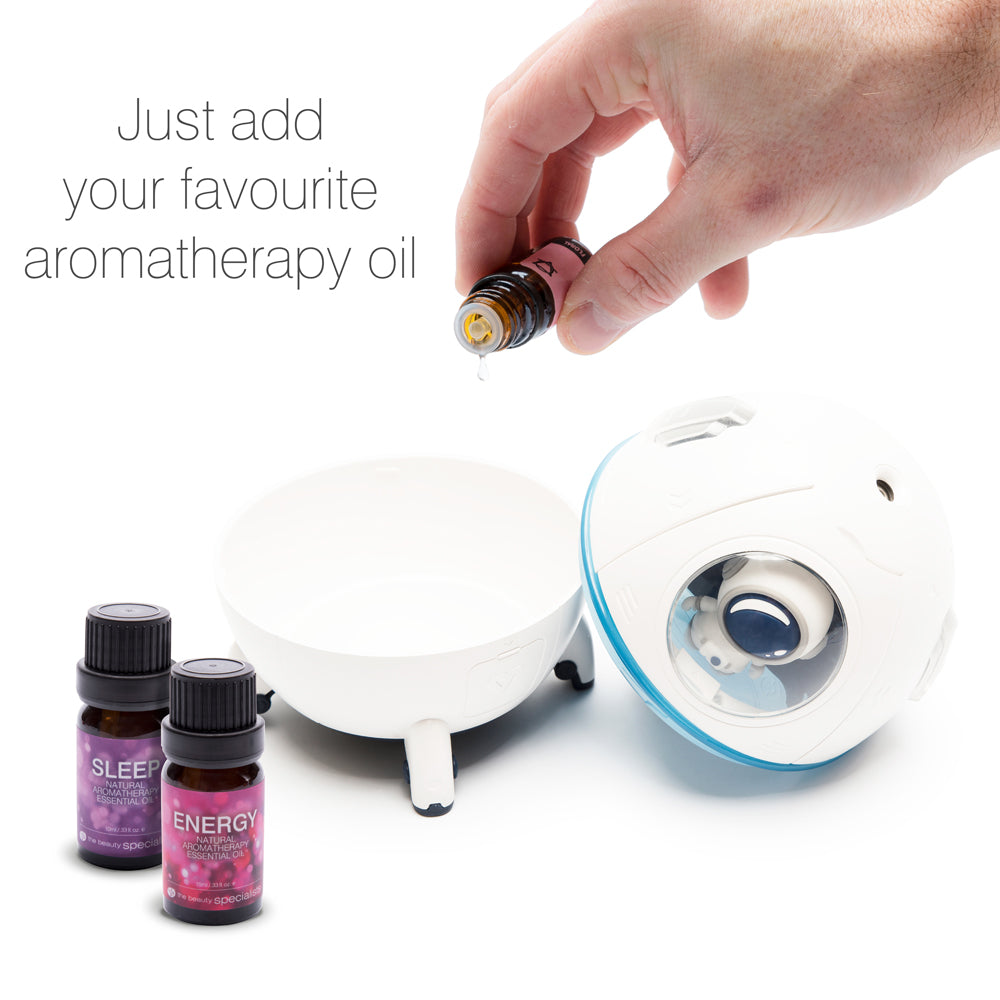 A hand adds aromatherapy oil to a white Rio Spaceship Explorer Childs Essential Oil Diffuser, Humidifier and Night Light. Two SLEEP and ENERGY oil bottles are nearby. Text: “Just add your favourite aromatherapy oil.”.