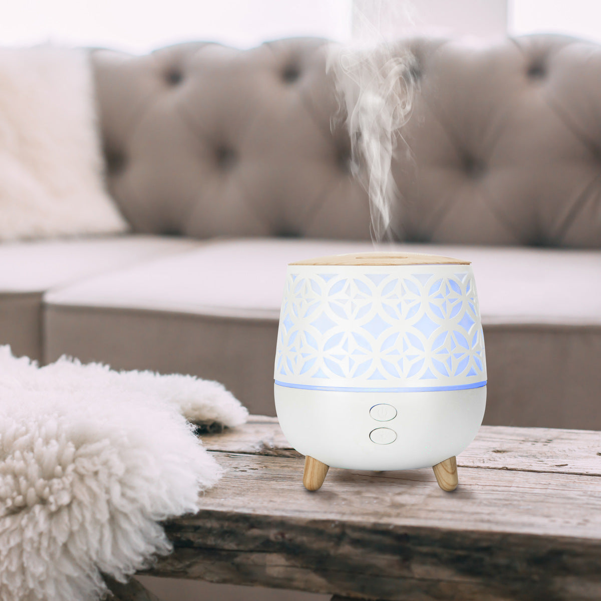 The Rio TALIA Aroma Diffuser, Humidifier & Night-Light emits vapor on a wooden table before a beige tufted sofa and white throw, creating a cozy living room while adding soothing light.