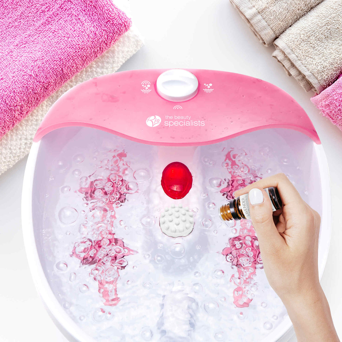 A hand pours essential oil into the Rio Soothing Waves Foot Bath Spa & Massager, its pink and white basin bubbling as towels sit nearby—a relaxing moment of pure indulgence.