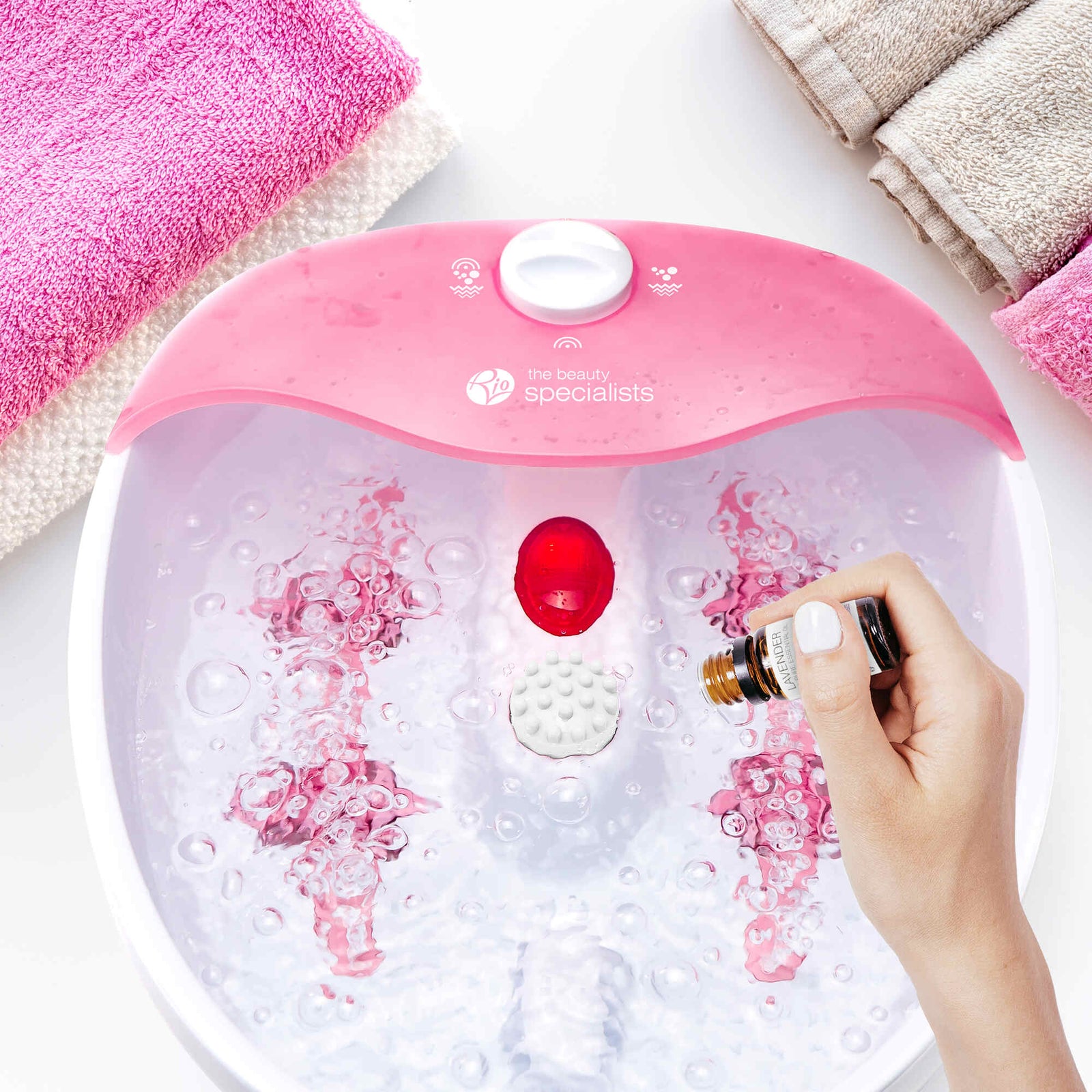 A hand pours essential oil into the Rio Soothing Waves Foot Bath Spa & Massager, its pink and white basin bubbling as towels sit nearby—a relaxing moment of pure indulgence.