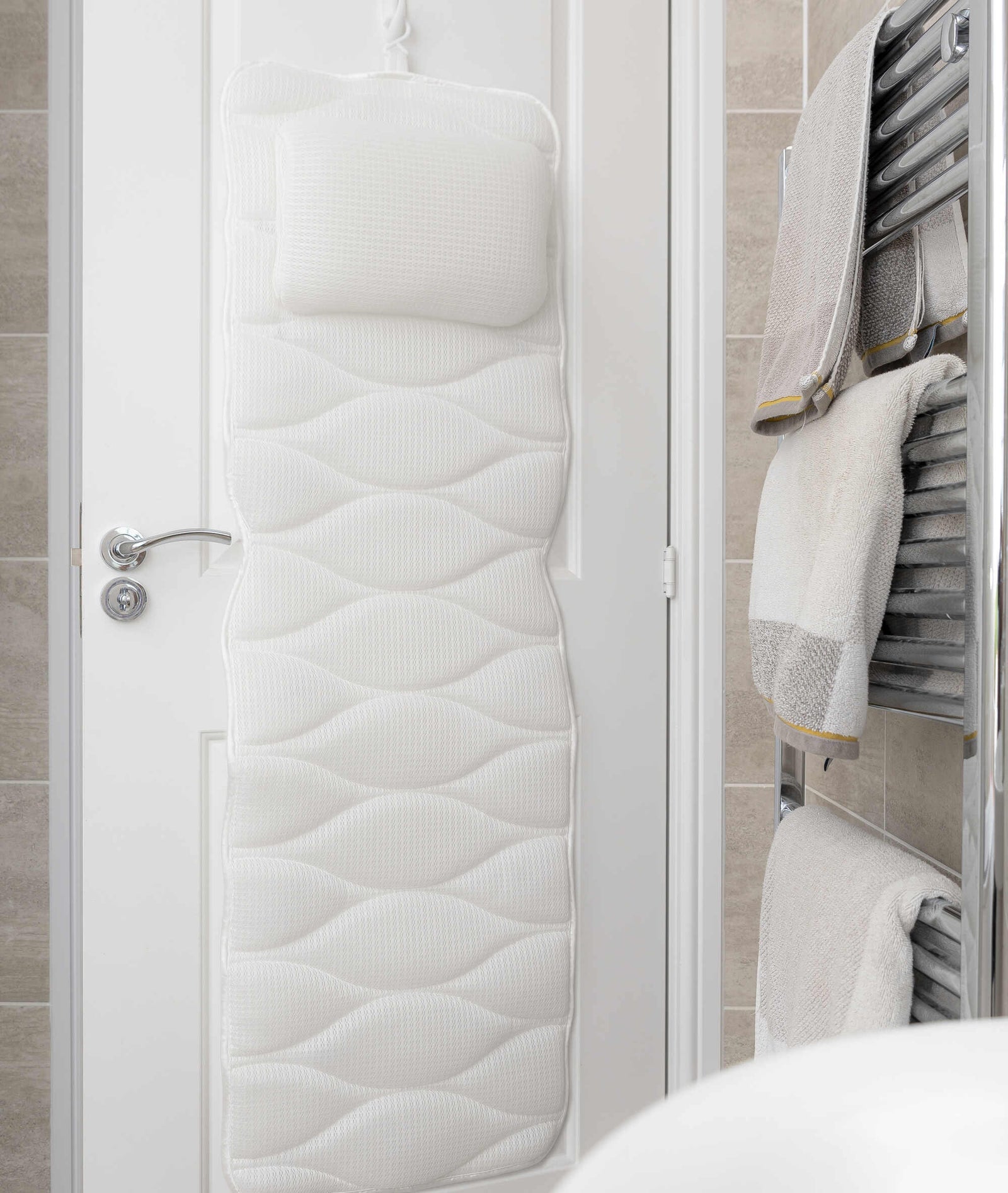 The Rio Luxury Bath Mat & Pillow, featuring a white padded design with headrest, hangs by the door near a heated towel rail with folded beige and white towels, creating an inviting space for bathing relaxation.