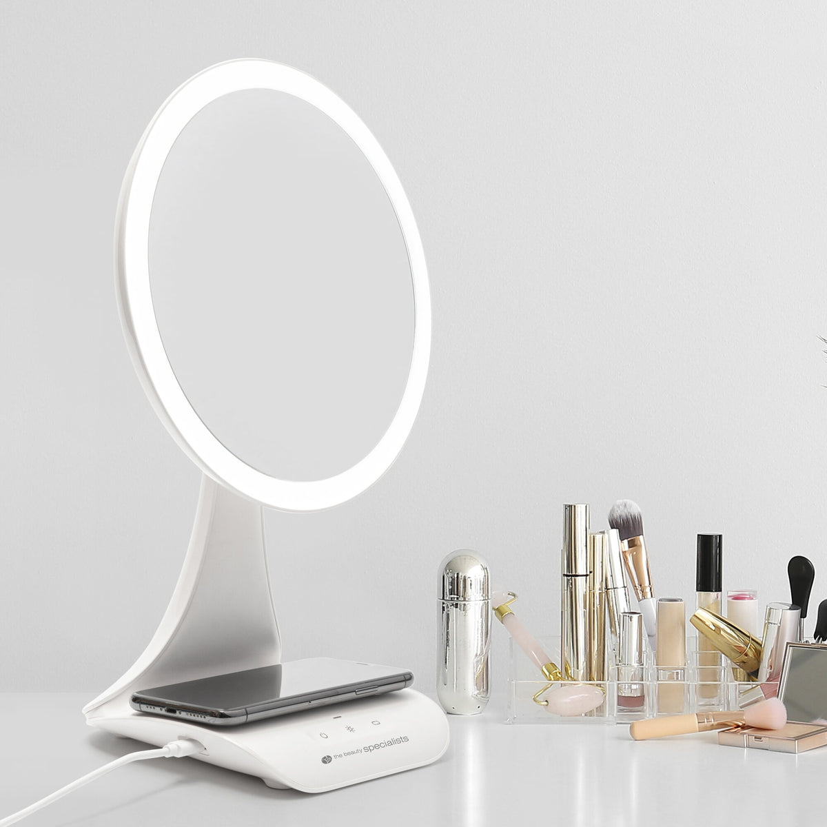 The Rio 5x magnification LED makeup mirror with a wireless charging station sits on a white surface, next to a clear organizer holding brushes and beauty products, against a plain white wall.