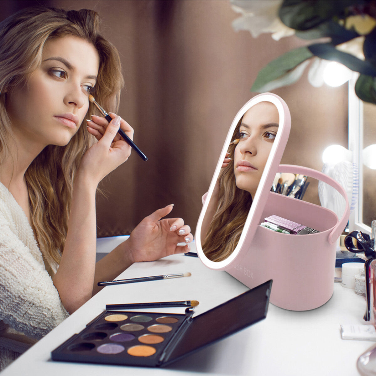 A woman in a bathrobe uses a brush to apply eyeshadow, looking into a lighted pink makeup mirror on her vanity. Her Rio Lush Box Vanity Case neatly holds her cosmetics and brushes.