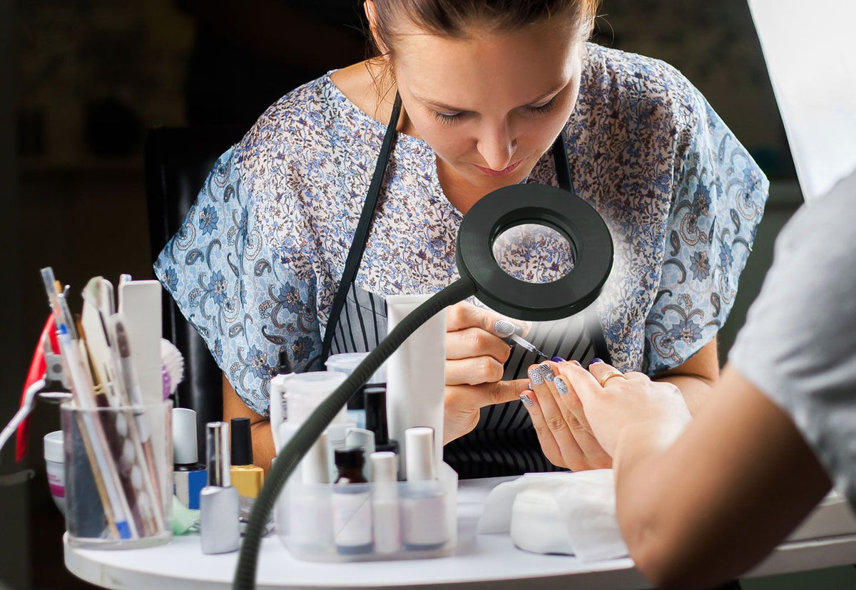 A nail technician in an apron applies polish to a clients toenails under the bright Rio Beauty Station LED Ring Light, with manicure tools and bottles arranged nearby.