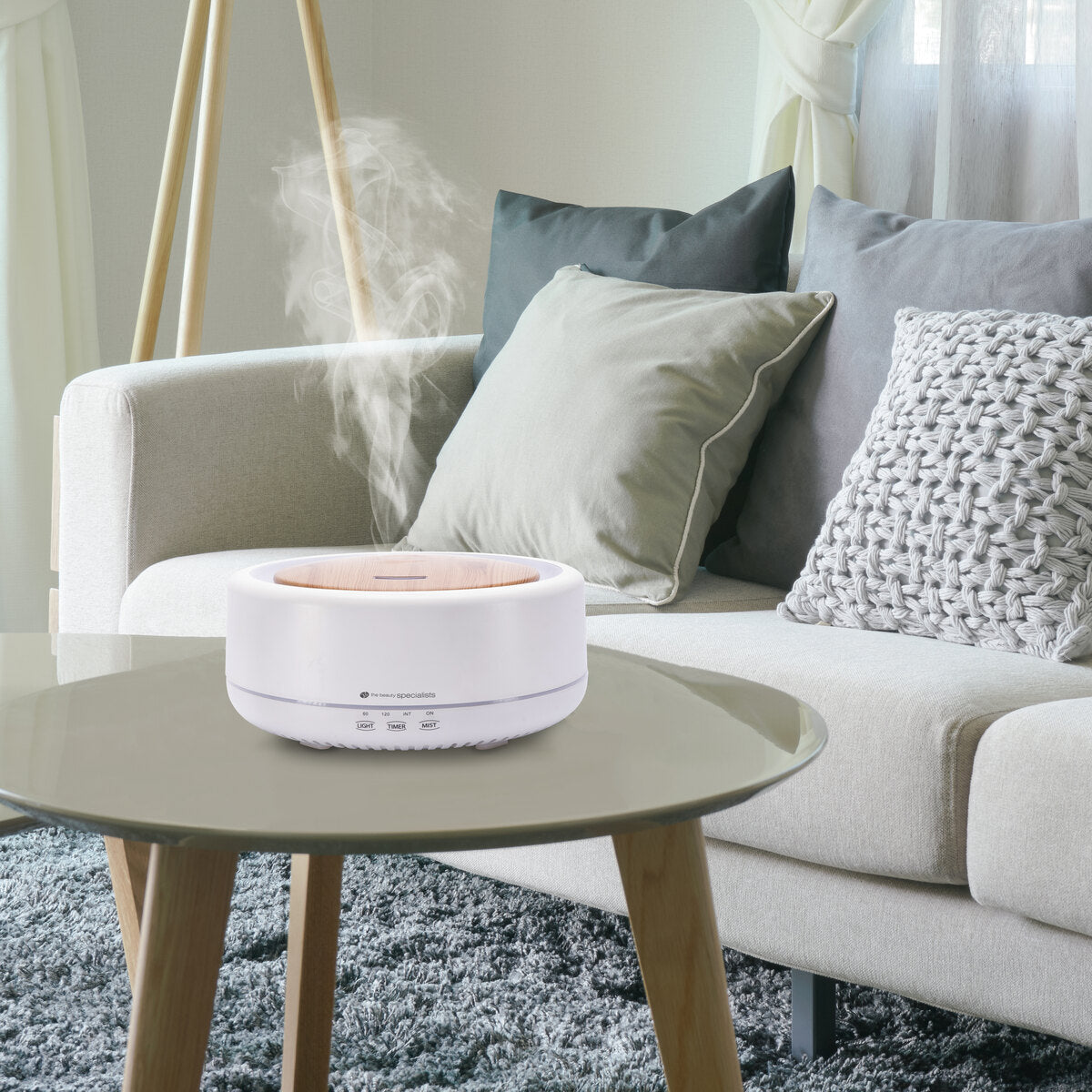 The Rio ENYA Aroma Diffuser, Humidifier & Night Light emits soothing mist on a glass coffee table in front of a light gray sofa with cushions, enhancing the relaxing ambiance of the cozy living room.