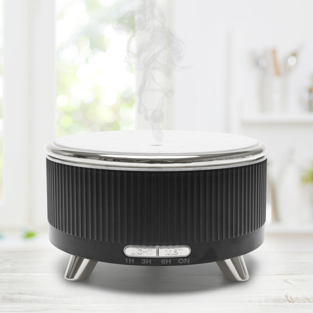 The Rio SONAS Aroma Diffuser, Humidifier & Night-Light in black and silver adds moisture to a bright kitchen, sitting on a light surface with blurred kitchen items and sunlight streaming through a window in the background.