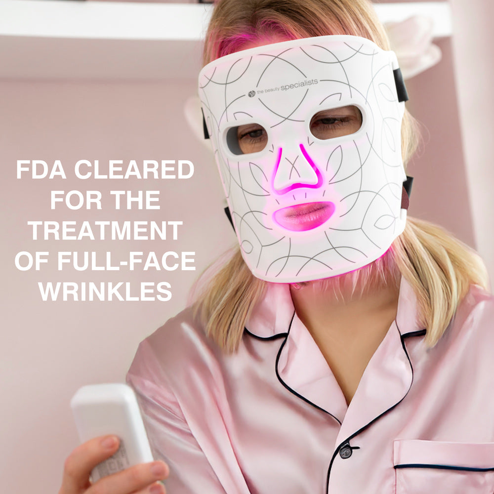 A woman in pink satin pajamas uses the Rio LED Face Mask by Rio, which emits pink light. Bold text: FDA CLEARED FOR THE TREATMENT OF FULL-FACE WRINKLES. She holds a controller, enjoying anti-ageing light therapy at home.