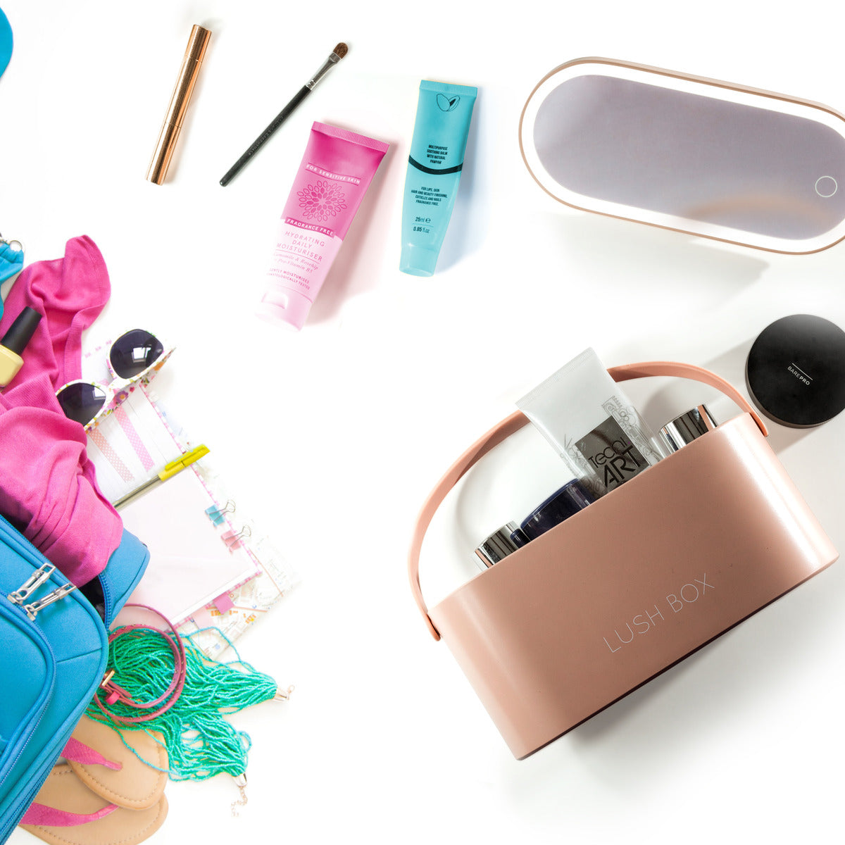 Flat lay of travel essentials: a blue suitcase, pink clothes, sunglasses, notebooks, skincare and makeup items, plus the Rio Lush Box Vanity Case—your perfect beauty companion—all neatly arranged on a white background.