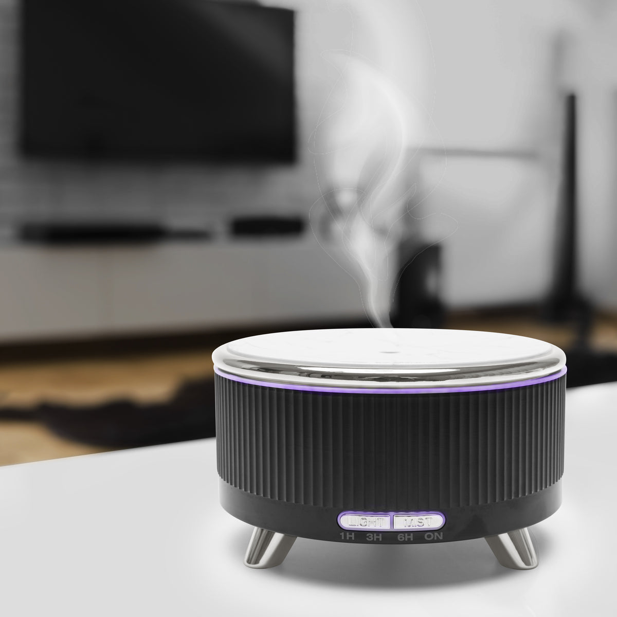 The Rio SONAS Aroma Diffuser, Humidifier & Night-Light in black and silver emits visible mist and soft mood lighting as it sits on a white surface in a stylish living room with blurred TV and furniture in the background.