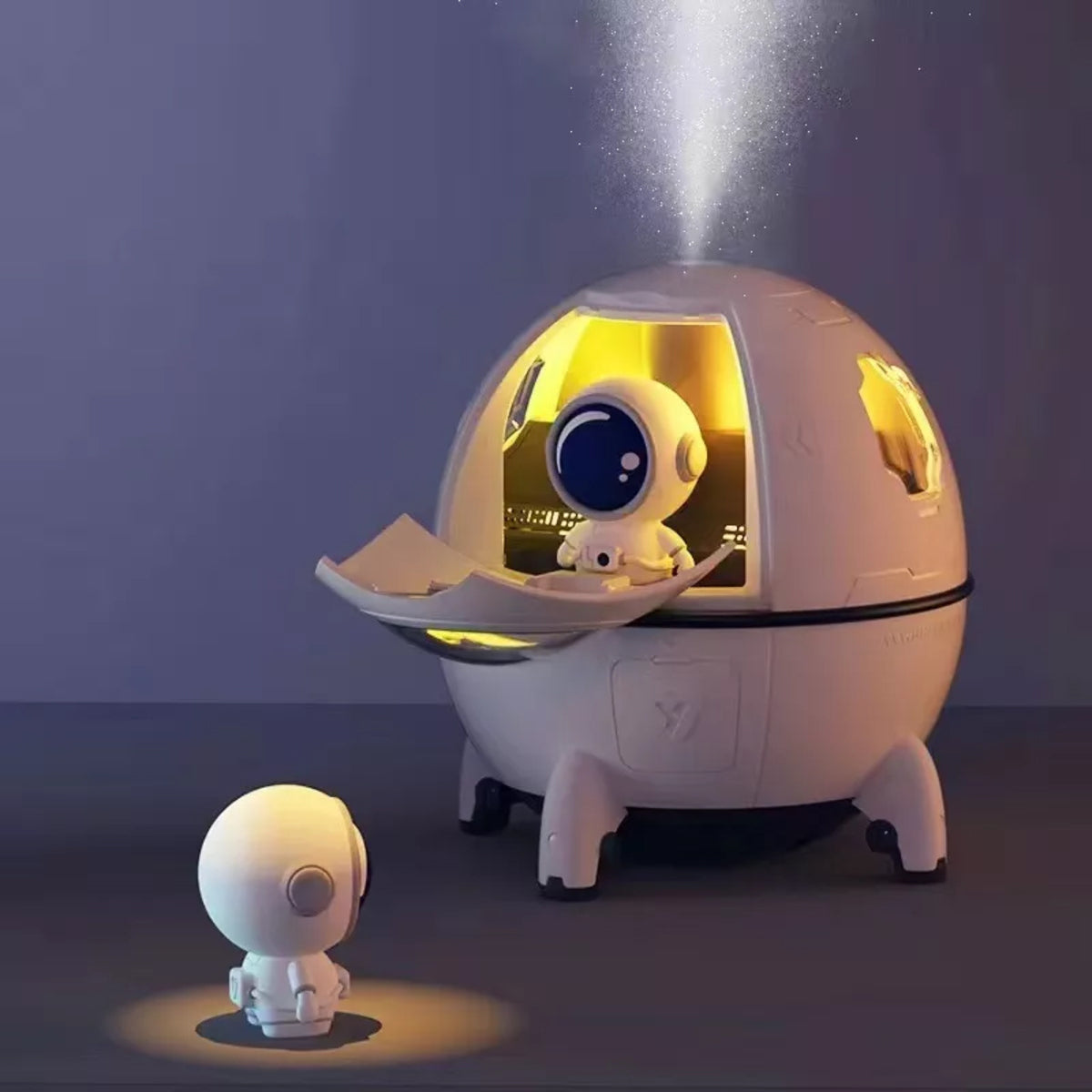 Two adorable robots interact: one stands on the floor, while the other sits inside the Rio Spaceship Explorer Childs Essential Oil Diffuser, Humidifier and Night Light. A warm yellow beam shines from the top of this fun spaceship-themed device.