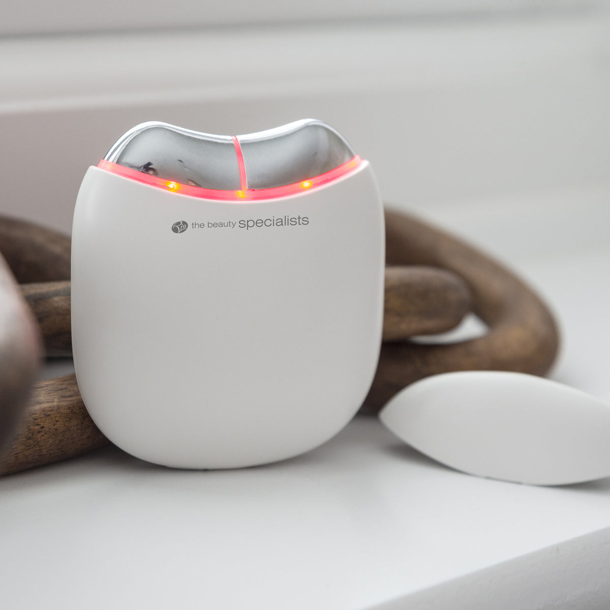 A compact, white Rio Face Lift & Tone Facial Toner with red indicator lights for skin toning rests on a white surface beside a decorative wooden object. The device is branded the beauty specialists.