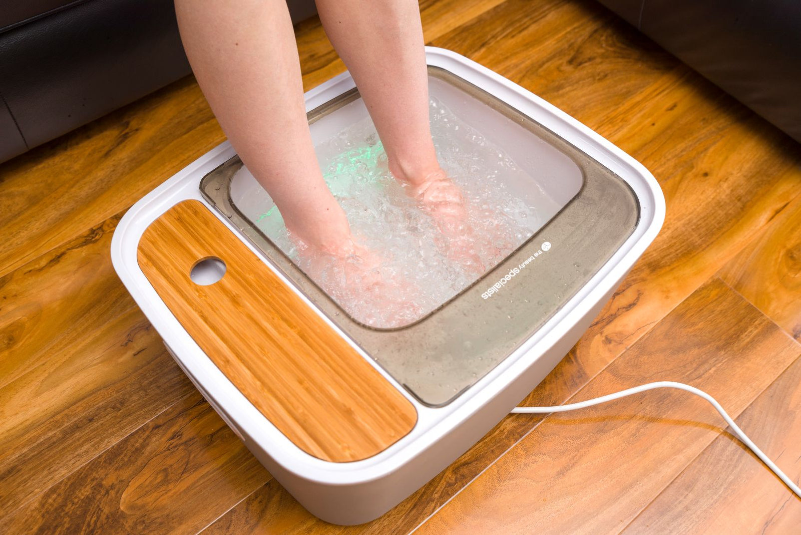 A persons feet soak in the Rio Scandinavian Jacuzzi Foot Spa Bath on a wooden floor, featuring a bamboo accent and visible power cord for an ultimate luxury foot spa pedicure.