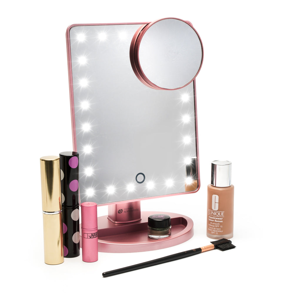The Rio Rose Gold 24 LED Makeup Mirror, rectangular and pink, includes a mini removable 10X magnifying mirror. It’s shown with cosmetic essentials like mascara, lipstick, foundation, an eyebrow brush, and more beauty products.