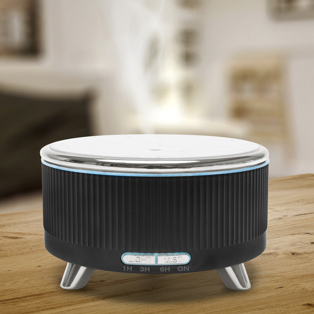 The Rio SONAS Aroma Diffuser, Humidifier & Night-Light in black and silver rests on a wooden table in a softly lit room, with front control buttons for timer, mood lighting, and light settings.