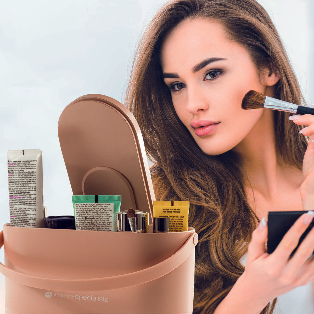 A woman with long brown hair applies makeup with a brush while looking in a mirror. In front of her is the Rio Lush Box Vanity Case, filled with beauty products and brushes.