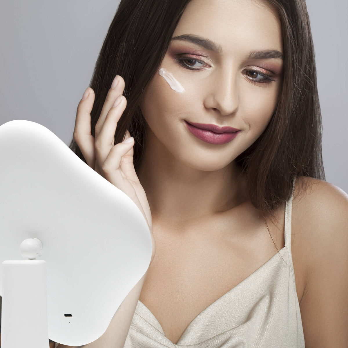 A young woman with long dark hair, wearing a light sleeveless top, smiles gently as she applies cream to her cheek while looking into the Rio Illuminated Full Size Beauty Mirror.