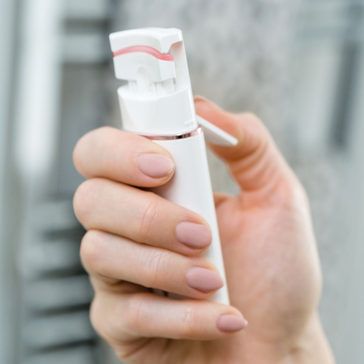 A hand with manicured nails and curled lashes holds the Rio WOW Lash Heated Eyelash Curler, poised for use against a blurred grey background.