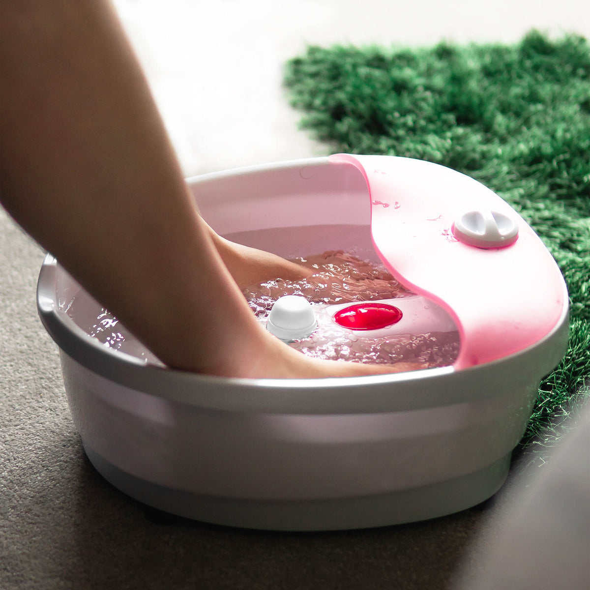 A person enjoys a relaxing soak with the Rio Soothing Waves Foot Bath Spa & Massager, feet immersed in its pink and white basin filled with water, placed on a carpeted floor near a green rug—pure at-home indulgence.