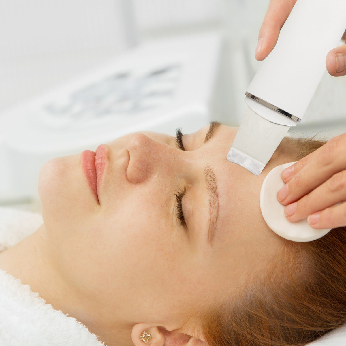 A woman with closed eyes receives a relaxing facial at a spa as a specialist gently exfoliates her forehead with the Rio Ultrasonic Skin Cleanser while holding a cotton pad.