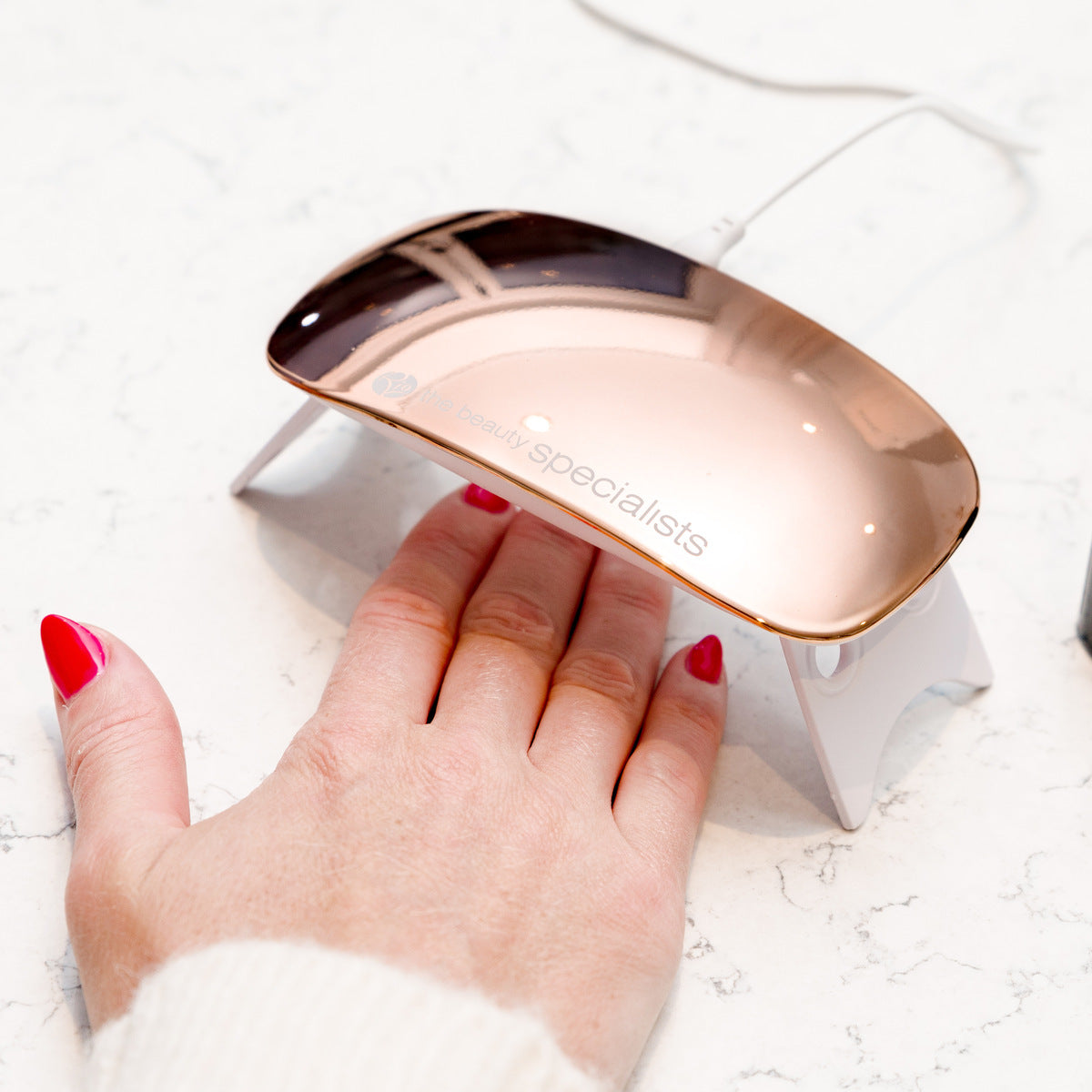 A hand with red 14 Day Gel Polish Nails by Rio is placed under a compact rose gold UV lamp on a white counter for a professional, long-lasting manicure.
