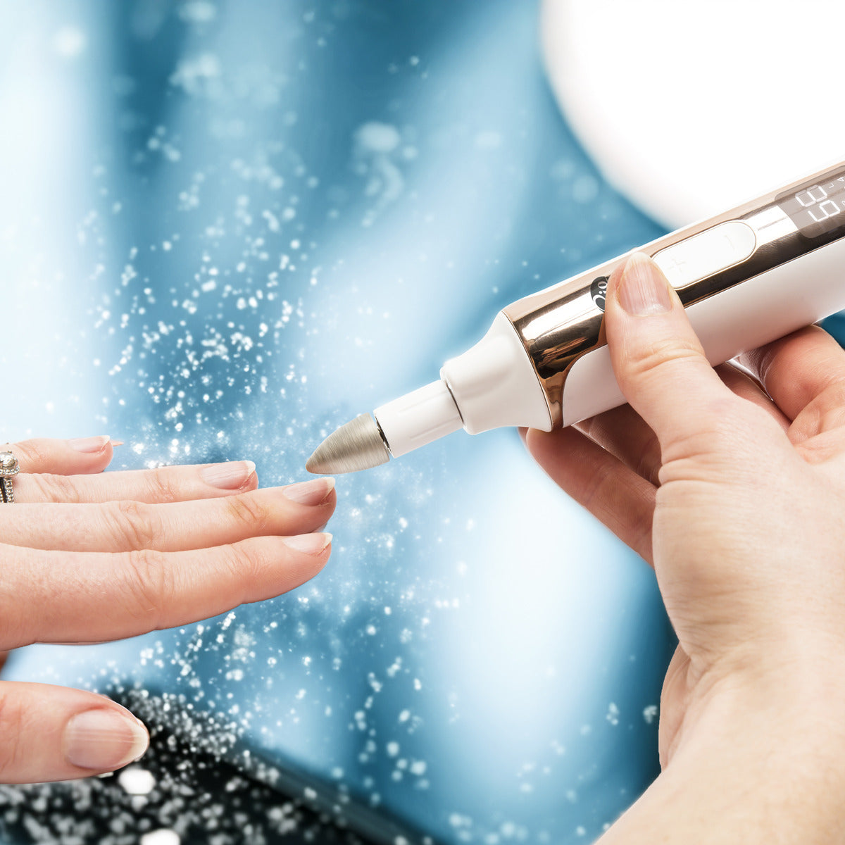 A close-up of a hand having nails filed with the Rio Precision Pro Electric Nail File, capturing precision and nail dust in the air against a blurred blue background—ideal for showcasing professional nail care.