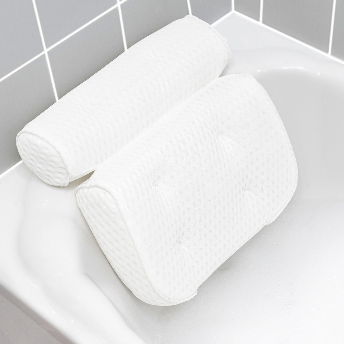 The Rio Luxury Bath Spa Pillow, featuring a white textured design with two plush cushions, rests on the edge of a white bathtub beside gray and white tiled walls.