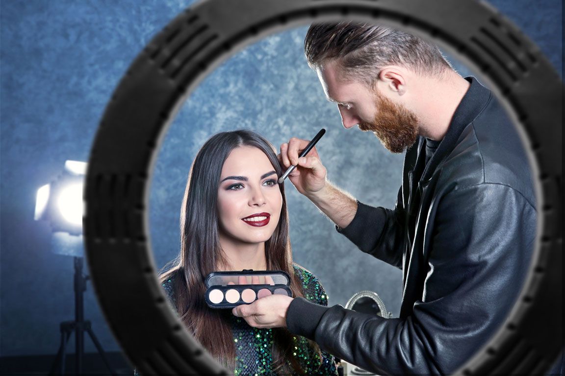 A makeup artist uses the Rio Professional Makeup & Vlogging 18-inch Dimmable LED Ring Light to apply eyeshadow to a smiling woman with long dark hair, reflecting in a circular mirror against a blue background.