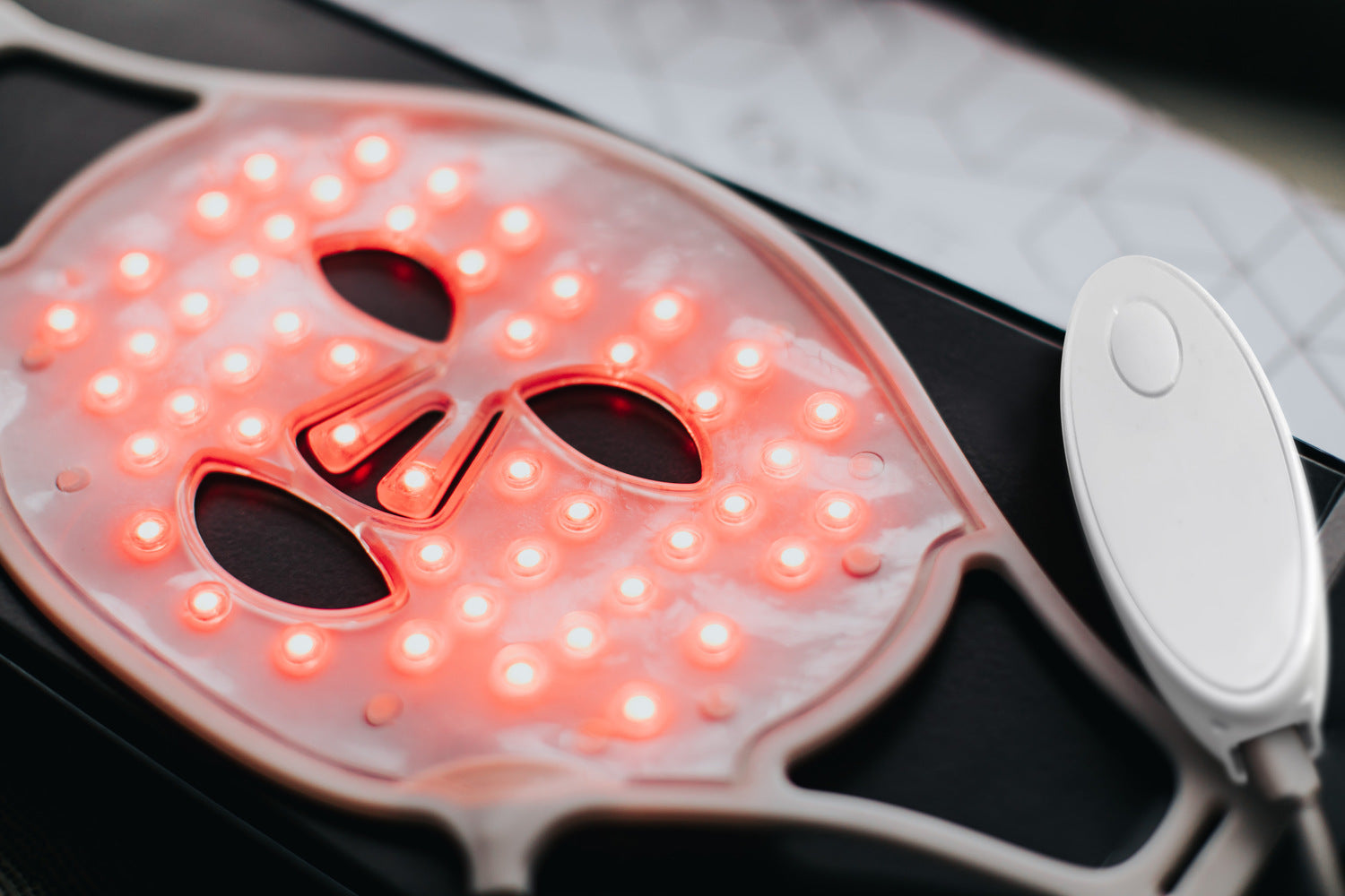 A close-up of the Rio faceLITE beauty boosting LED face mask glowing red for light therapy skincare. Designed to energize and target aging skin, it features eye, nose, and mouth cutouts and connects to a white controller.