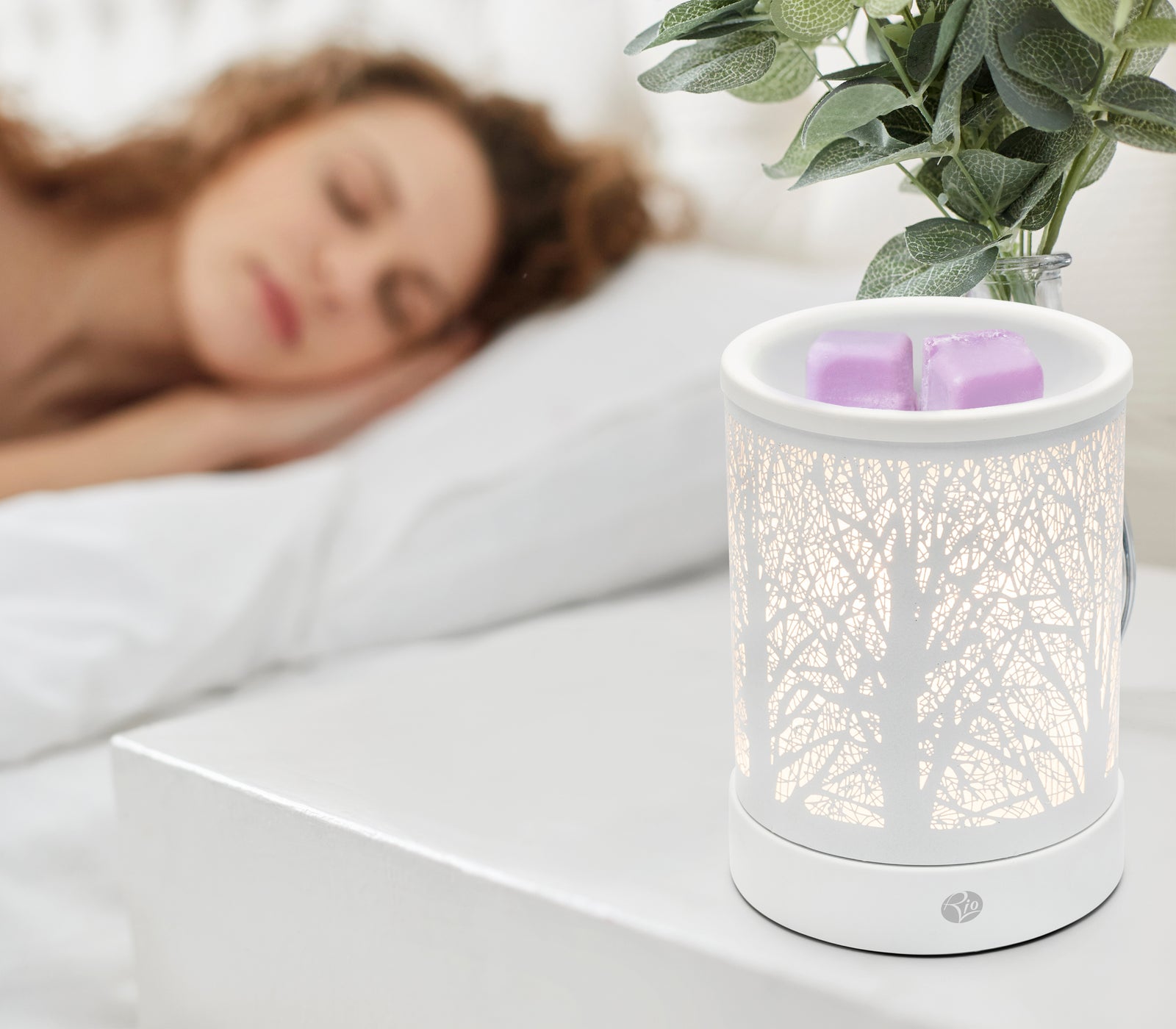 A woman sleeps on a white bed beside a nightstand with the Rio FORA Wax Melt Burner & Aroma Diffuser Lamp glowing softly, holding two purple wax melts and a green potted plant, letting her relax in style.