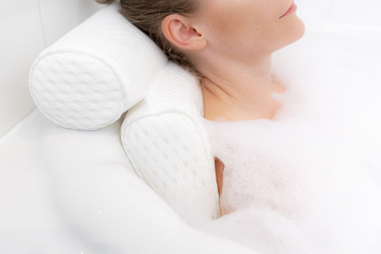 A person relaxes in a bubble bath, resting their head and shoulders on the white, textured Rio Luxury Bath Spa Pillow. Only the lower face and upper body are visible.