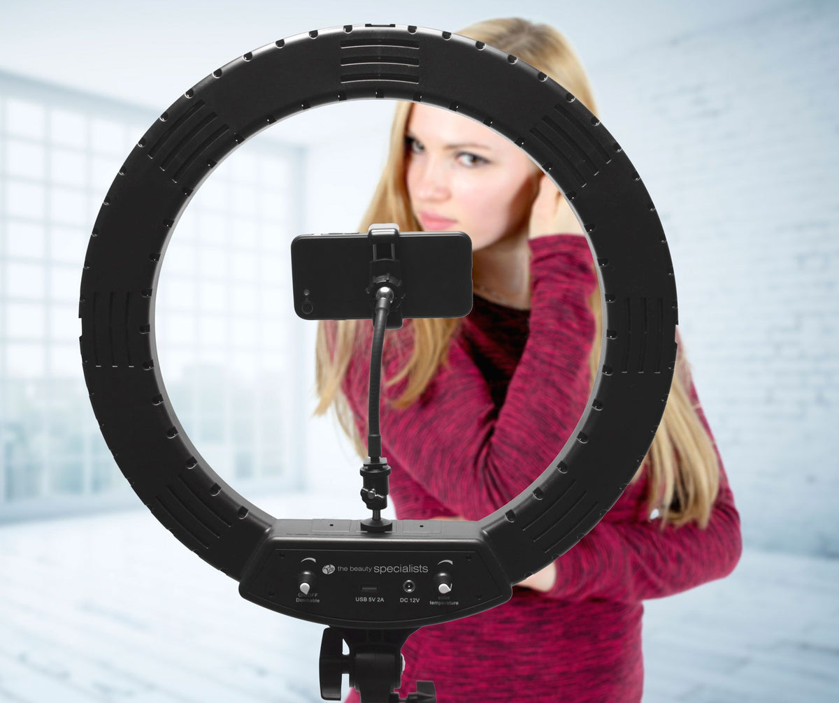 A woman in a maroon top adjusts her hair while facing her smartphone mounted in the center of the Rio Professional Makeup & Vlogging 18-inch (45cm) Dimmable LED Ring Light, enjoying ideal lighting in a modern, sunlit room.
