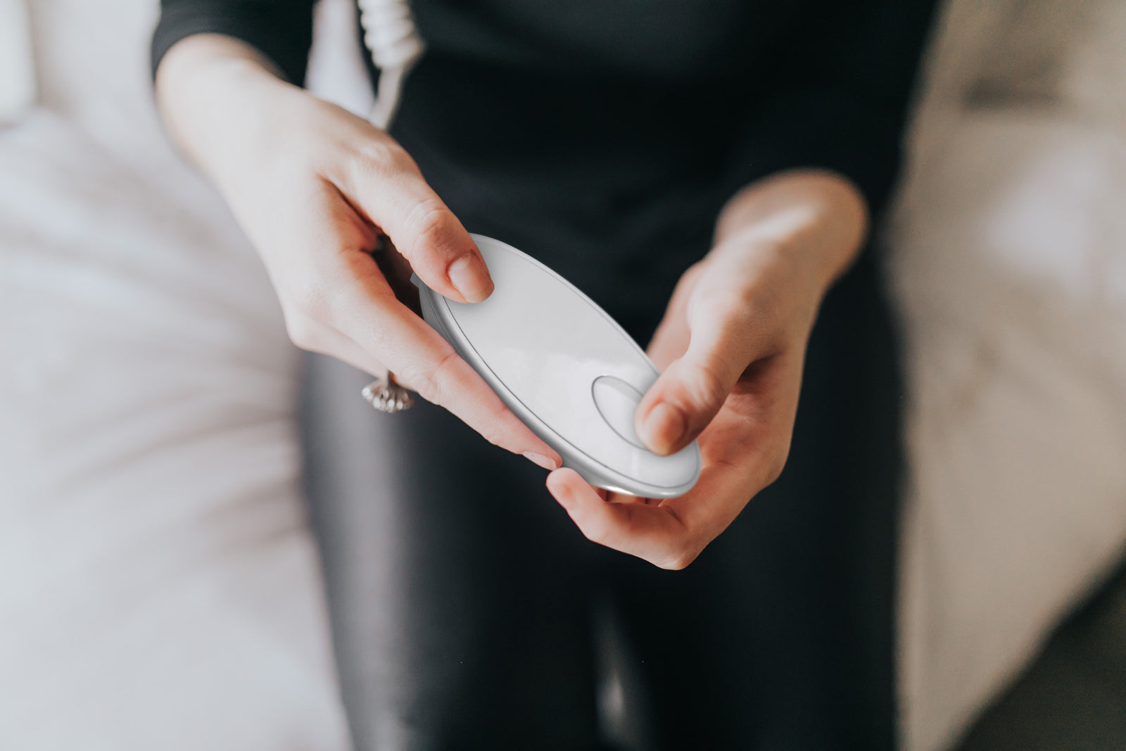 A person in black clothing holds the Rio faceLITE beauty boosting LED face mask, a white oval device with a single button, while sitting on a bed—designed to energize and rejuvenate ageing skin.