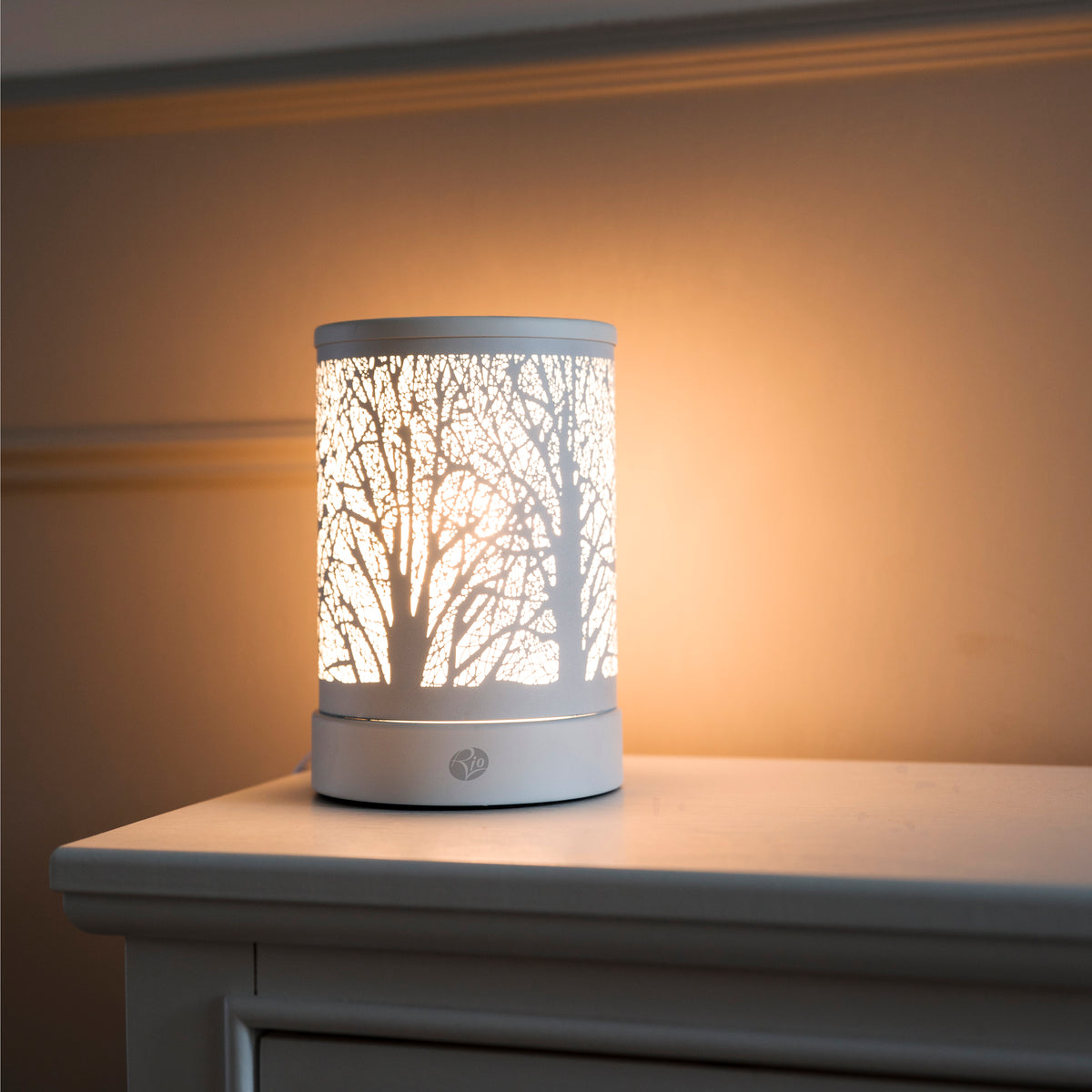 The FORA Wax Melt Burner & Aroma Diffuser Lamp by Rio features a tree branch design, casting a soft, warm glow on any white dresser—perfect for relaxing in style against a beige wall.