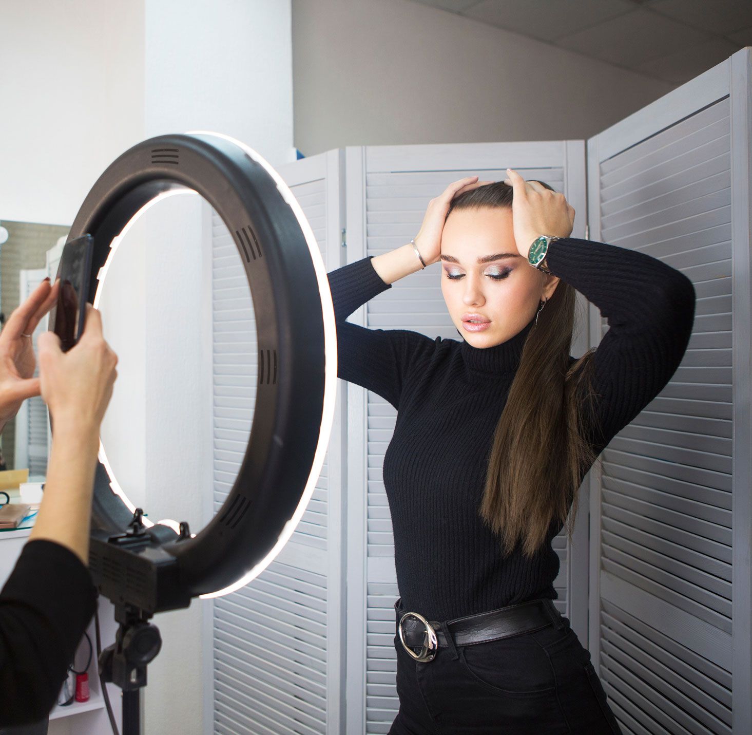A woman poses in front of the Rio Professional Makeup & Vlogging 18-inch Dimmable LED Ring Light, enjoying perfect lighting as someone takes her photo with a smartphone. Gray folding screens are visible in the background.