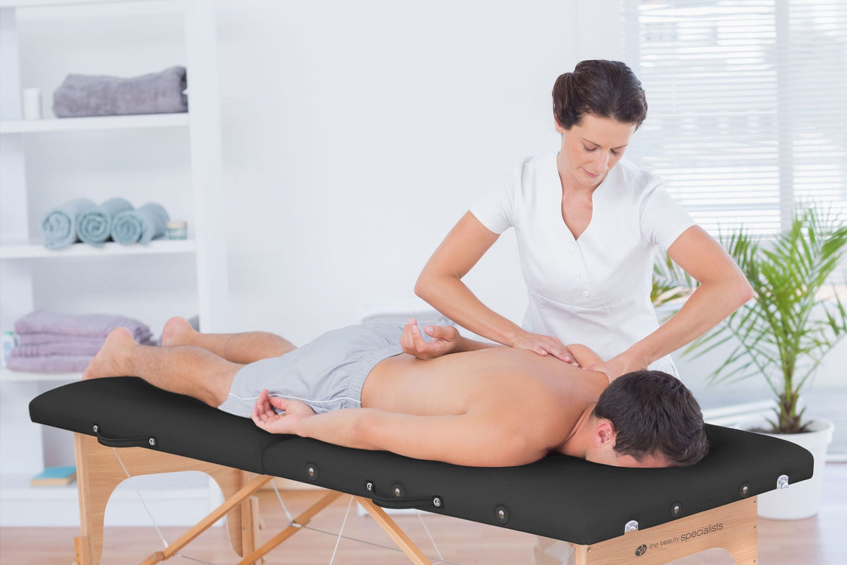 A massage therapist in a white uniform gives a back massage to a man lying face-down on the Rio Professional Ultra-Light Portable Massage Table in a bright, relaxing room with towels and plants in the background.