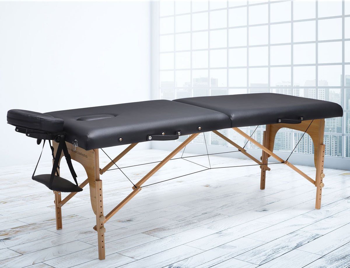 The Rio Professional Ultra-Light Portable Massage Table, in black with wooden legs, is set in a bright room. It features an adjustable face cradle and armrest for added comfort during professional massage sessions.