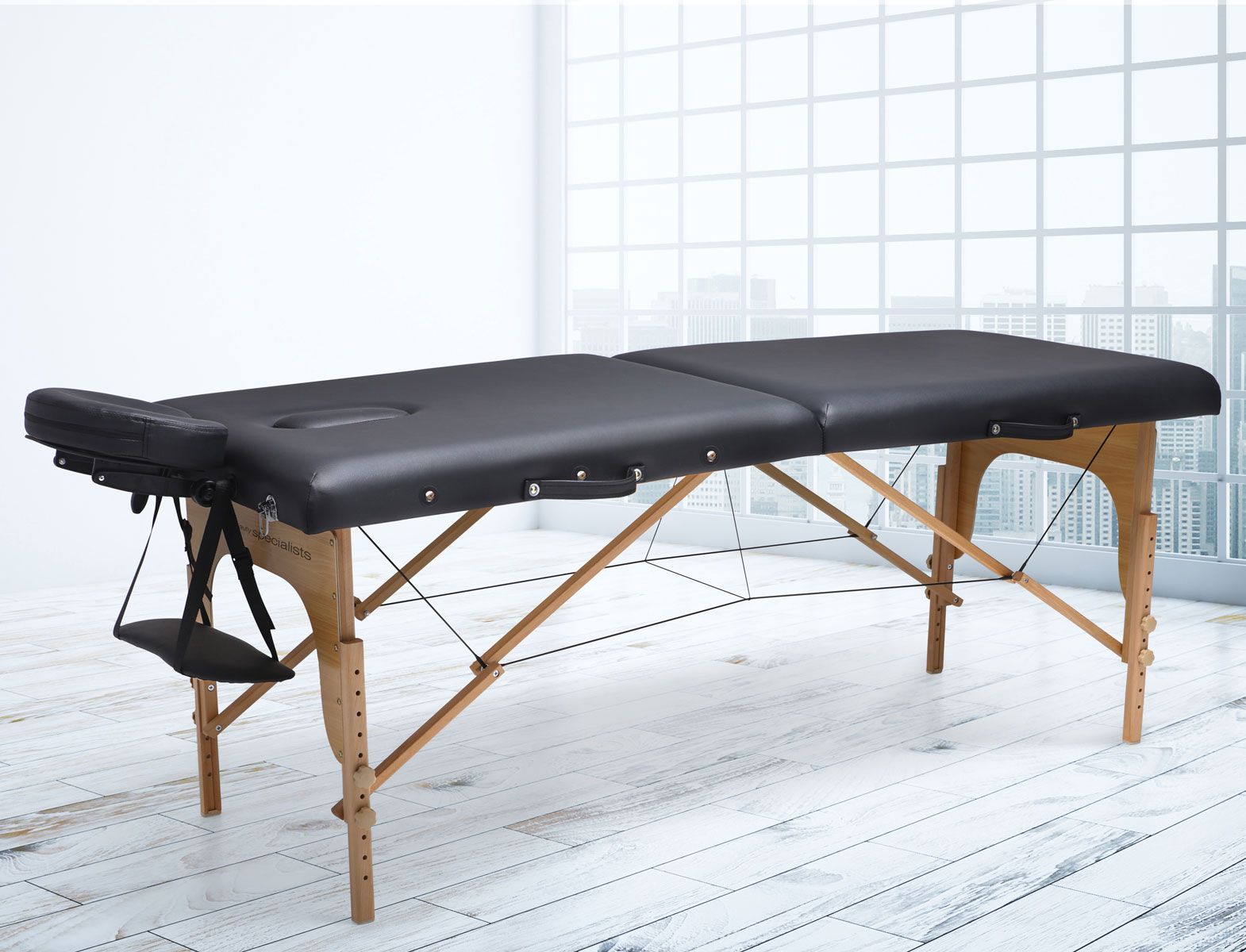 The Rio Professional Ultra-Light Portable Massage Table, in black with wooden legs, is set in a bright room. It features an adjustable face cradle and armrest for added comfort during professional massage sessions.